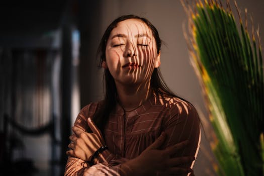 A woman embraces herself in serene indoor lighting with shadow play on her face.