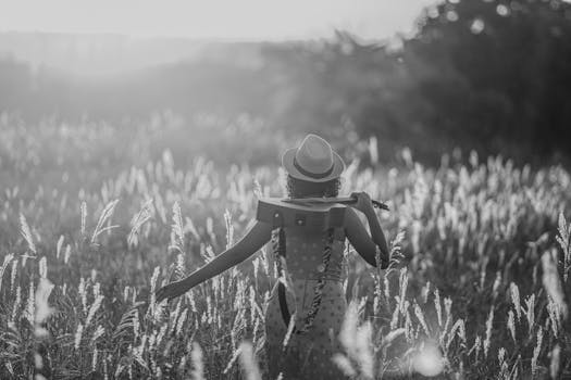 A woman in a sun hat walks through a grassy field with a guitar during sunset, capturing a serene moment.